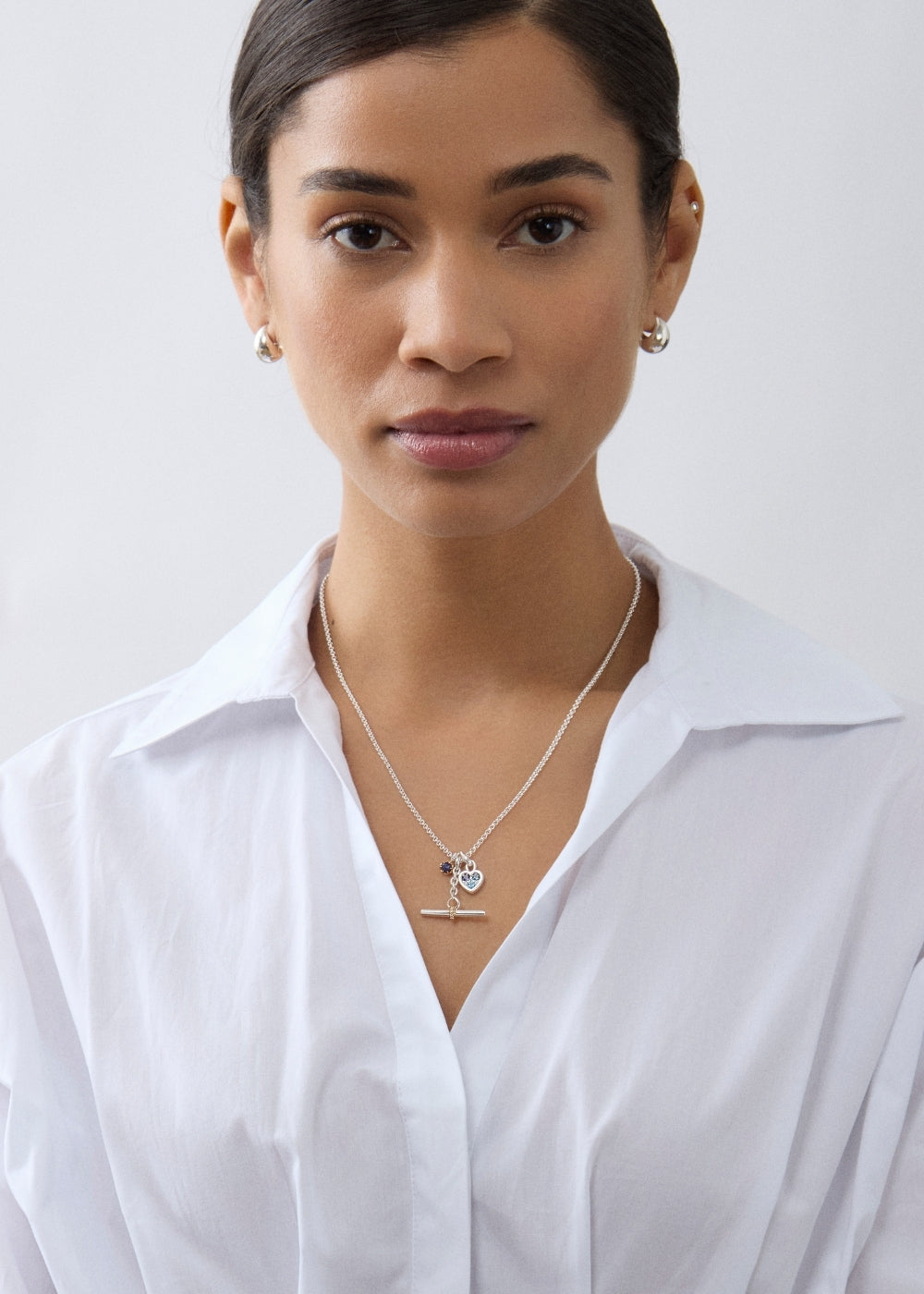 Layered necklaces with small gemstone charms and a T-bar, worn over a white shirt.